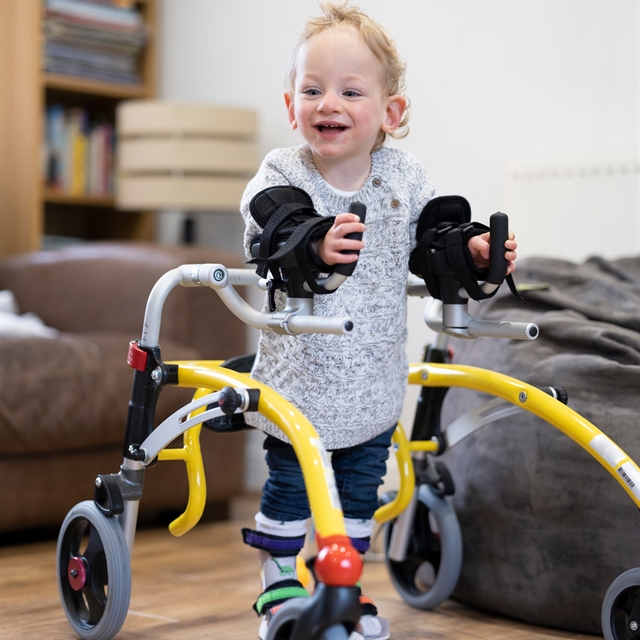 A young child with light hair is smiling and using a bright yellow pediatric walker for support.