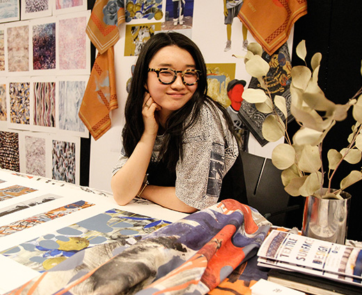 smiling student sitting at a desk surrounded by fabric and paper