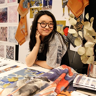smiling student sitting at a desk surrounded by fabric and paper